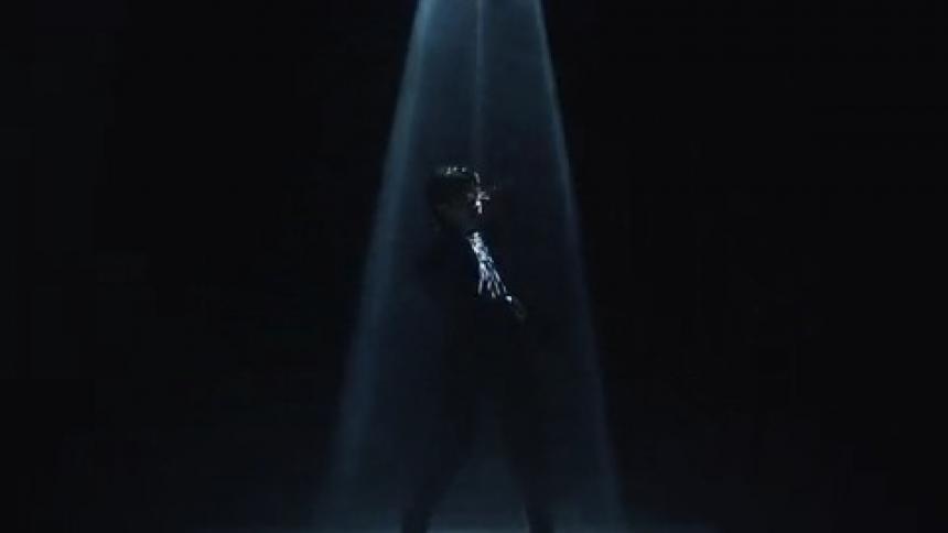 Woman in a black room poses and stands under a spot light that fades in and out