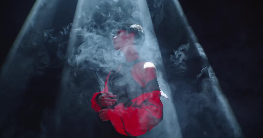 A model stands under a smoky light wearing a black dress with an orange coat around her shoulders