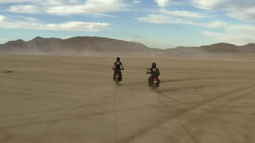 Two cyclists ride dirt bikes across the desert towards mountains on a cloudy day