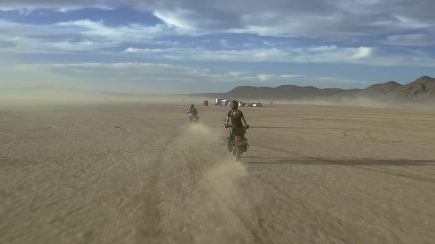 To cyclists riding dirt bikes across the desert on a cloudy day with mountains on the horizon