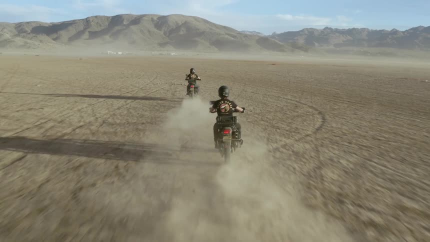 A pair of motorcyclists send up clouds of dust behind them as they ride through a barren flatland towards a distant mountain on a sunny day