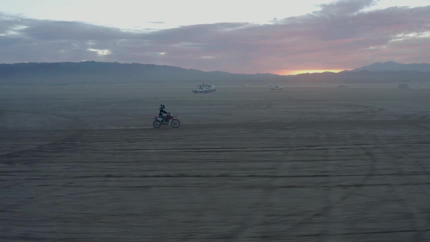 A person rides an offroad motorcycle across a vast barren flatland at sunset