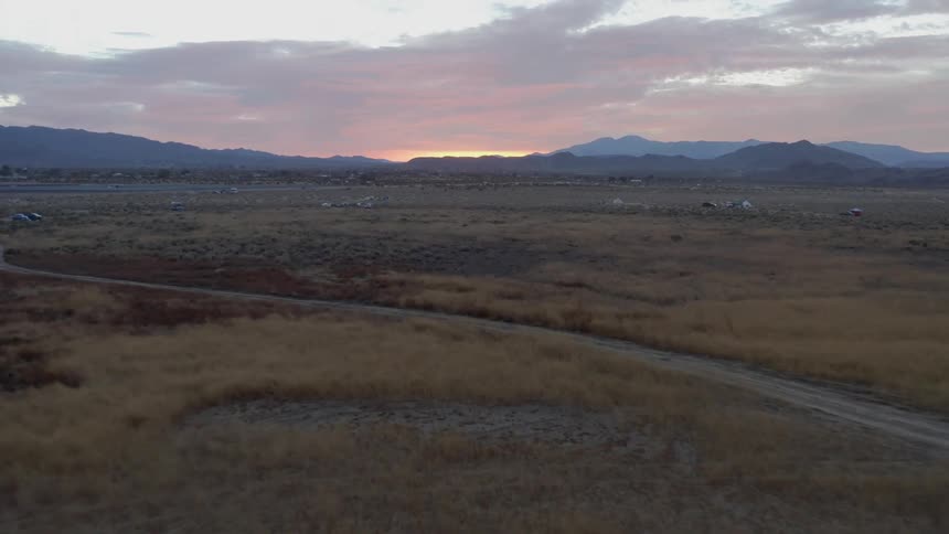 Vehicles are parked along a curved dirt road in a rural area with mountains in the distance