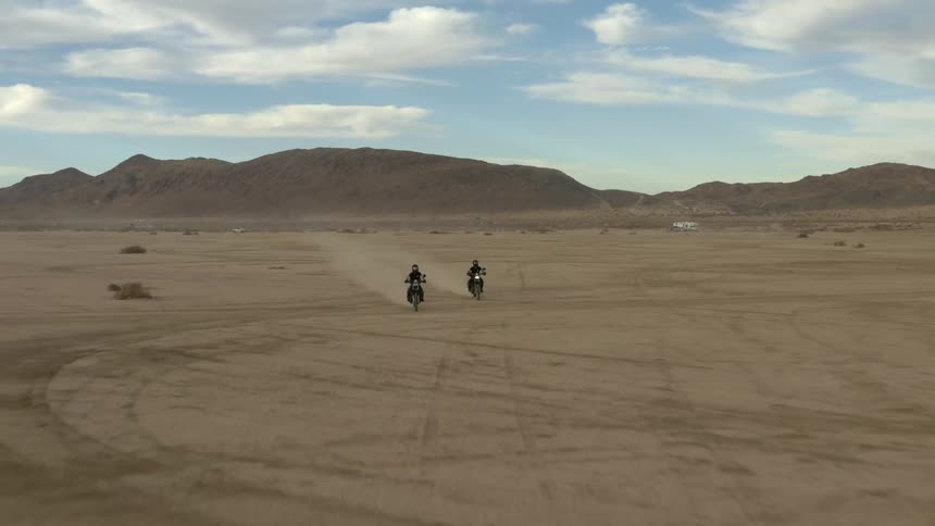 Two cyclists riding motorcycles across the desert with mountains behind them on a cloudy day