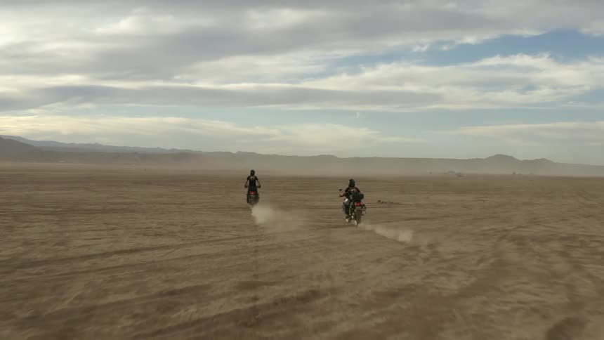 Two people with helmets riding motorcycles in the desert with the sky and clouds in the background