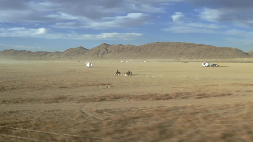 Two motorcycles ride in a desert passing other vehicles in front of a mountain
