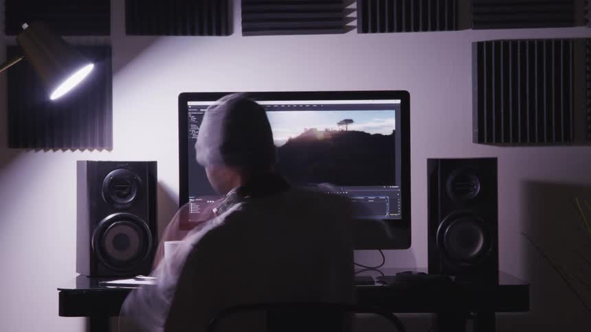 Time lapse of a man who walks over to a desk with a computer and sits down and edits videos with video editing software