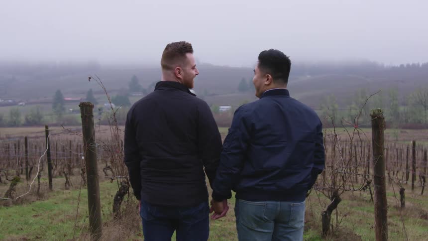 Two men holds and turn to smile at each other as they stand in front of crops on a farm