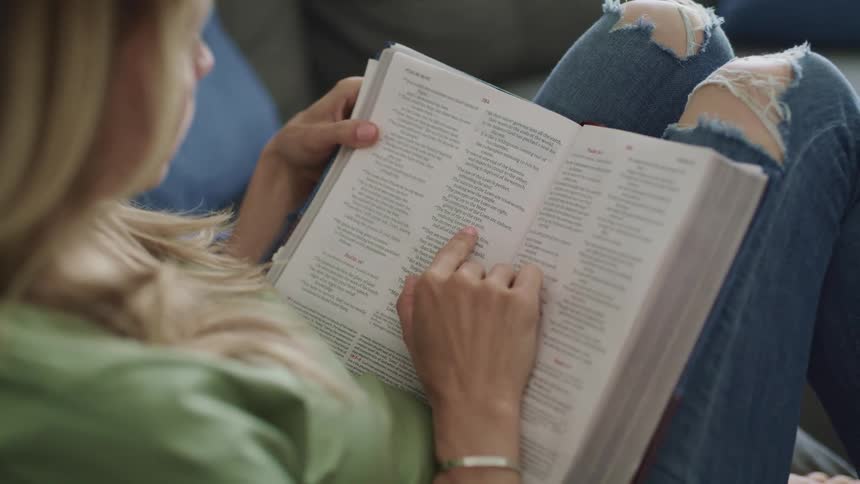 A woman sitting on a sofa runs her finger along a page of a bible as she reads