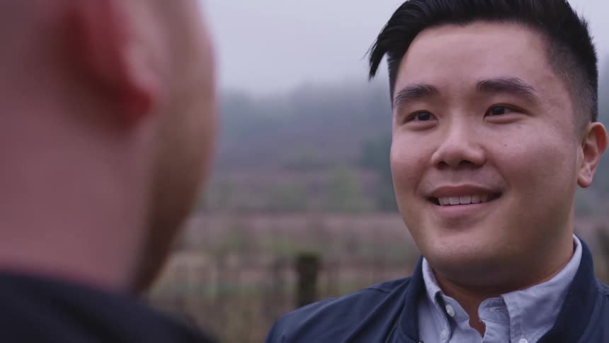 A man standing smiling and staring at another man with trees the sky and clouds in the background