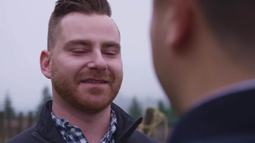 Man standing outdoors smiling in front of another man whos face is obscured