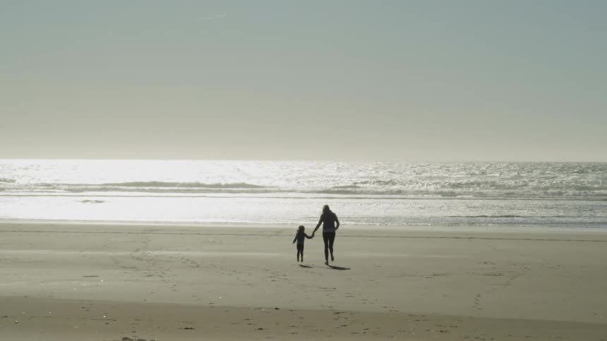 Mother and daughter run holding hands on the white sands of a beach near the ocean at day time