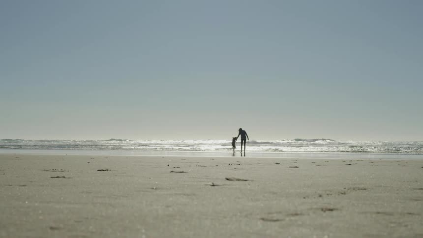 A woman holds the hand of a child on the beach as they stand at the edge of the water