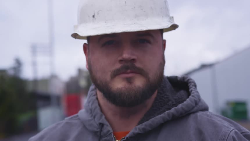 A bearded man wearing a construction hard hat looks directly into the camera