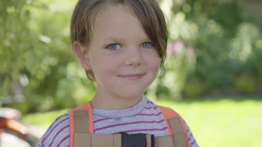 A young child smiles into the camera while standing outdoors during the day