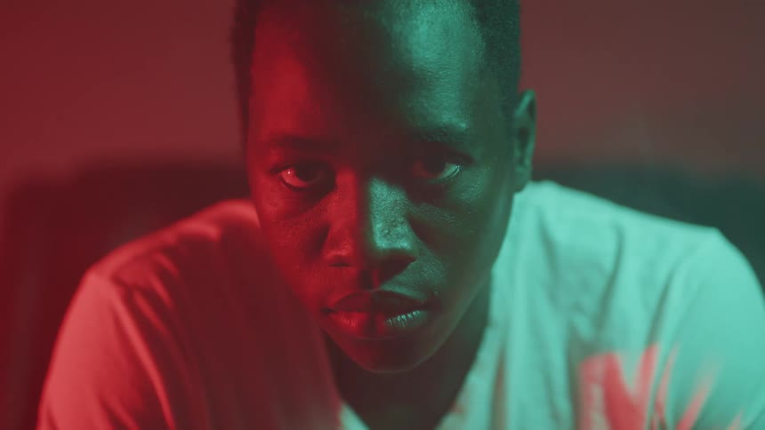 Close up of a young mans face sitting in a room under green and red lights