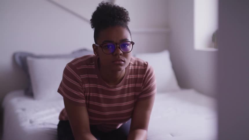 Young teenage woman sitting on a bed in the bedroom looks at the camera