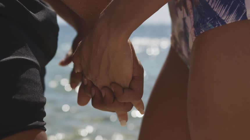 Two people face one another and hold each others hands while standing in front of a body of water