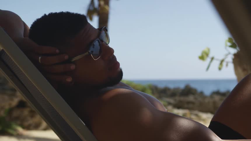 A black man wearing sunglasses lounges on a chair outdoors on a beach