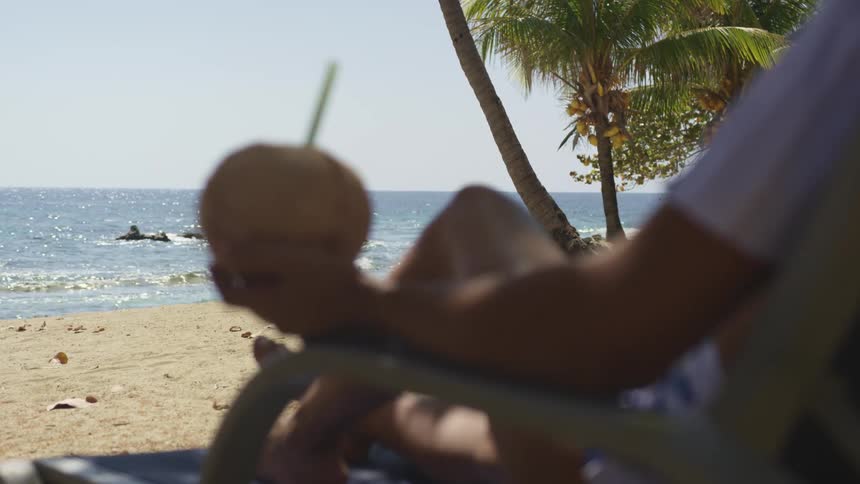 Man resting on a reclining chair in the beach holding a coconut cocktail