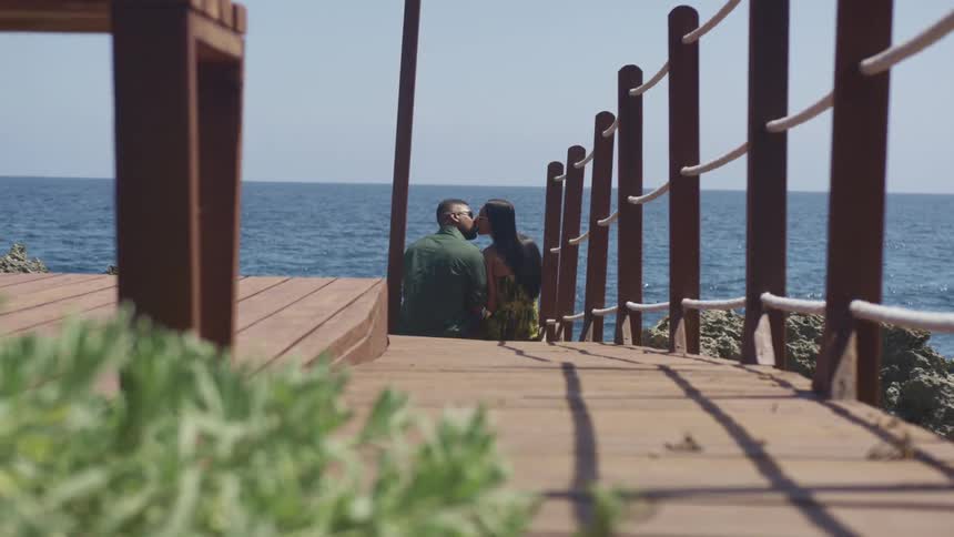 A black couple sit at the end of a dock overlooking water kissing on a sunny day