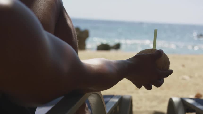 A man holding a coconut with a straw in it relaxes at a sunny beach