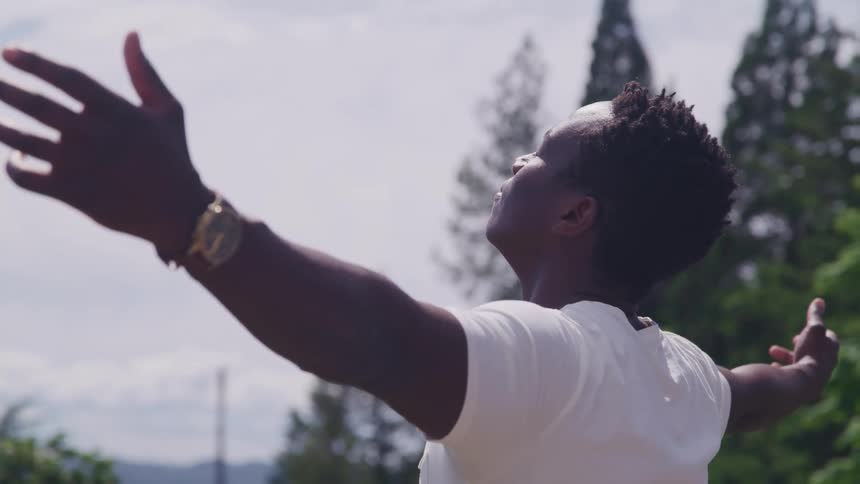 A black man wearing a white t shirt and wrist watch stands outdoors and stretches out his arms and closes his eyes