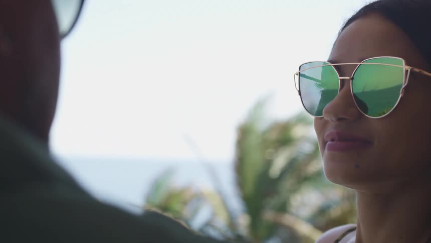 A man and woman sitting outdoors on a sunny day look at one another through their sunglasses