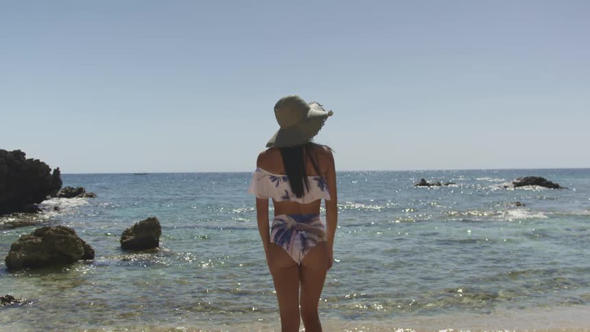 Young woman wearing swimsuit and sun hat stands on the sand of a tropical beach