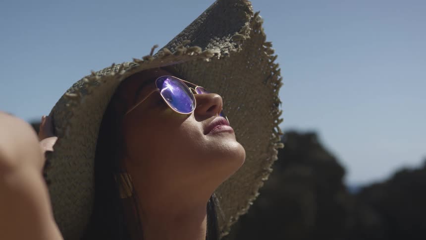A woman wearing printed tinted aviator sunglasses holds onto her straw hat as she looks up at the sun