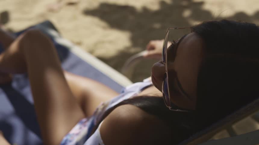 A woman in a tank top sits on a lounge chair on a beach in the shade while wearing sunglasses