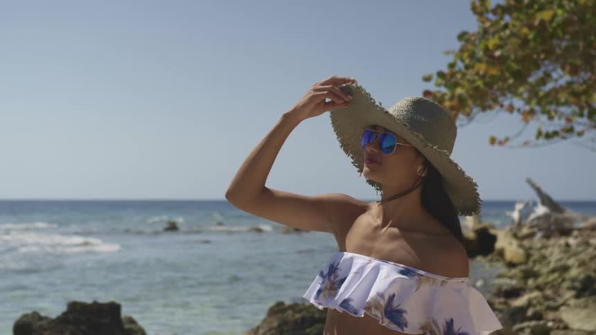 A woman stands outside near the ocean as she holds her sunhat because of a light breeze