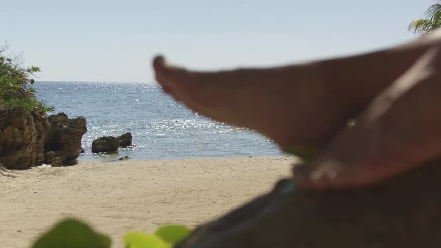 A persons feet laying down on a tree in a beach with sand the ocean the sky and clouds in the background