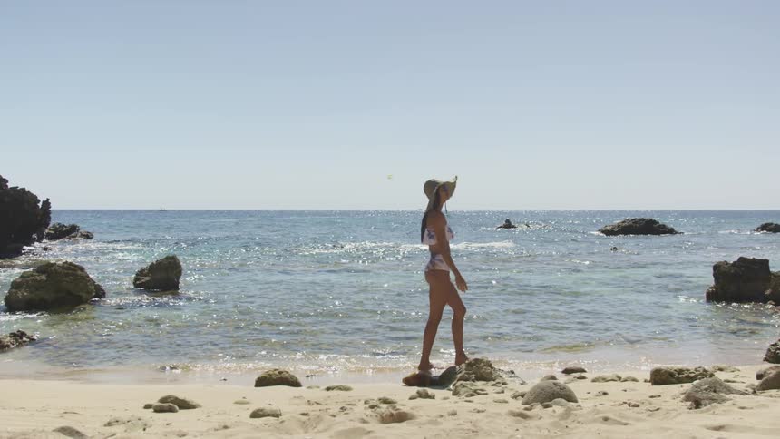 A woman wearing a swimsuit and hat walks along the beach on a sunny day