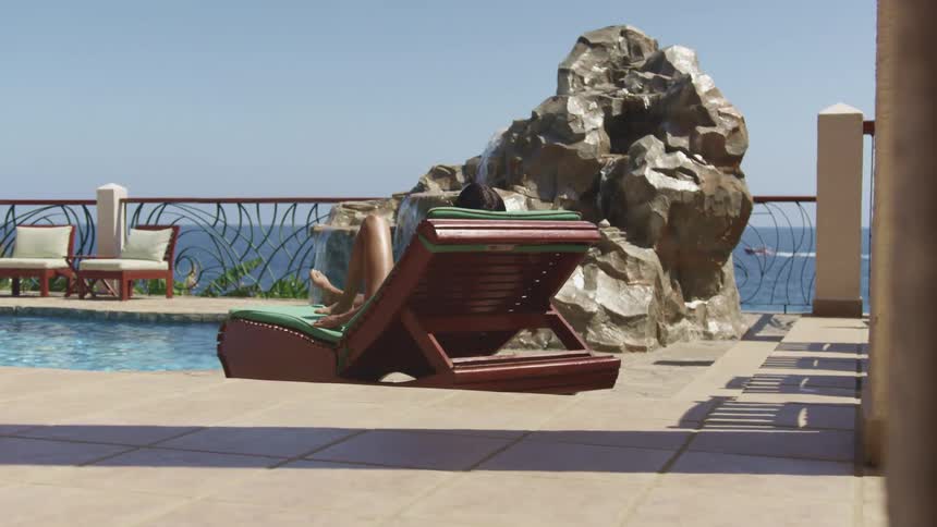 A woman relaxes as she reclines on chair outdoors on a sunny day next to a pool