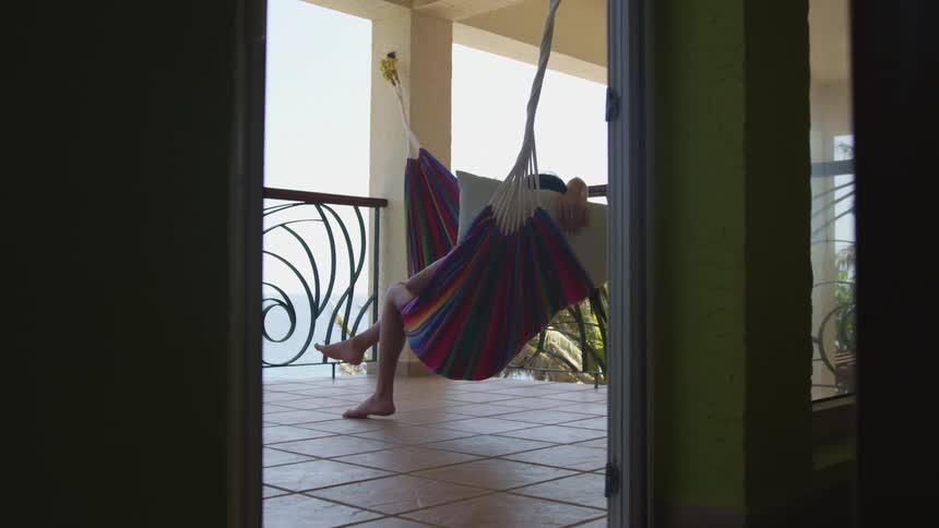 A woman relaxes just outside her room in a hammock on a balcony overlooking the blue ocean