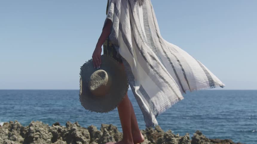 A barefoot woman holding a sunhat stands on the coast as her clothing billows in the wind on a sunny day