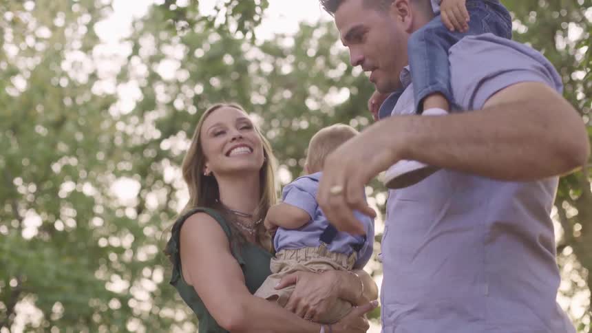A man with a small child sitting on his shoulders kisses a baby being held by a woman on the cheek while standing outdoors during the day