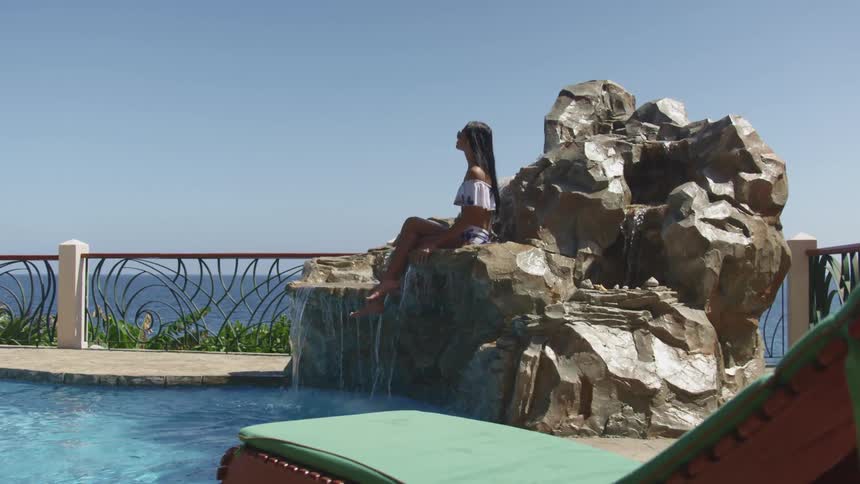 A woman sits on a decorative waterfall flowing into a pool which is located near the ocean