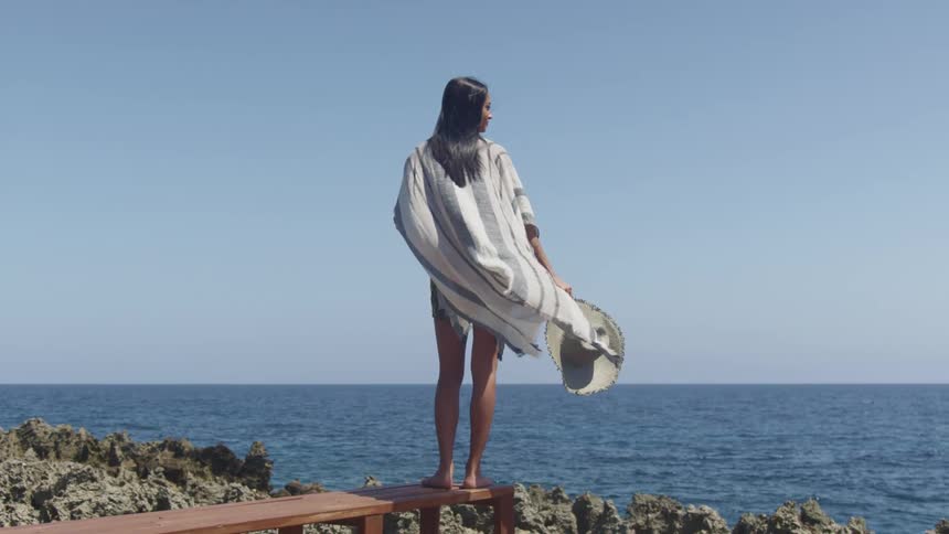 A woman with a hat and scarf blowing in the wind standing by a coast with the ocean sky and clouds in the background