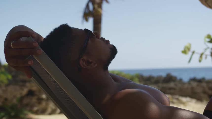 A black man lies on a lounge chair on the beach wearing sunglasses