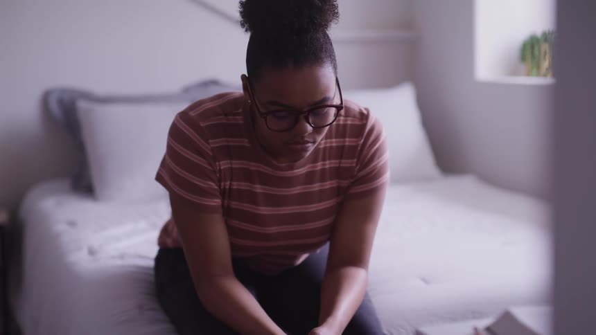 Young girl sits at the edge of a white bed in a bedroom and stares down at the floor sadly at daytime