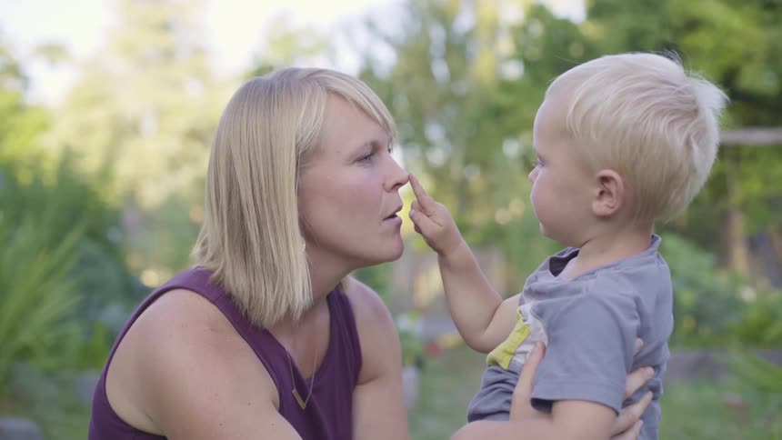 A woman kneeling before a small boy holds him in place and talks to him as he touches her face with his finger