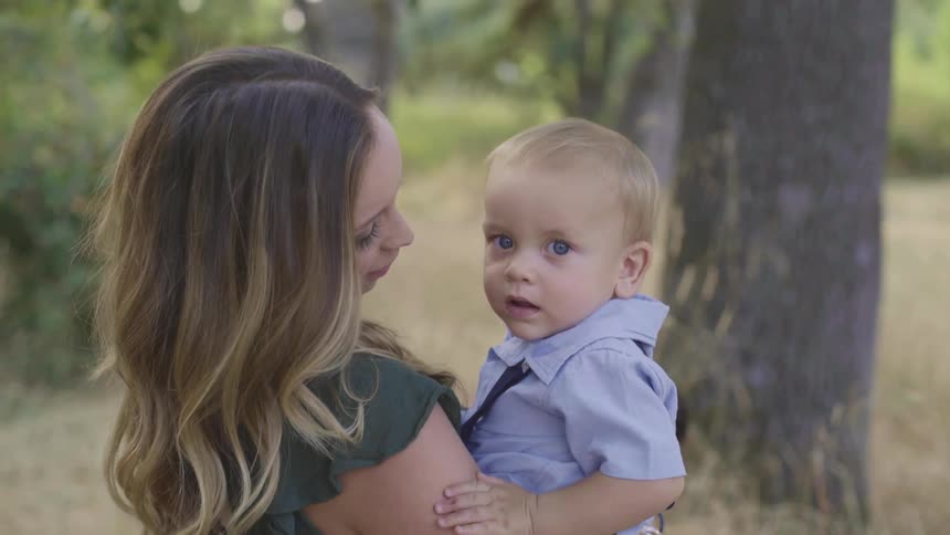 Mom holds her son in her arms standing outdoors near trees during the day