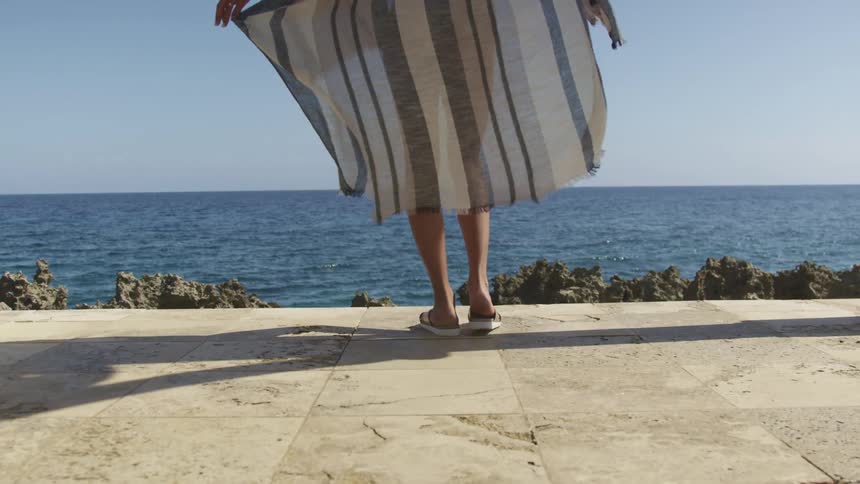 A woman wearing a striped caftan stands outdoors overlooking a body of water during the day