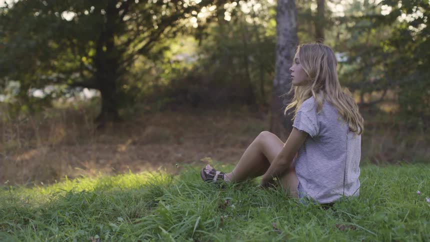 Girl with long hair wearing a dress sitting on the grass near the woods