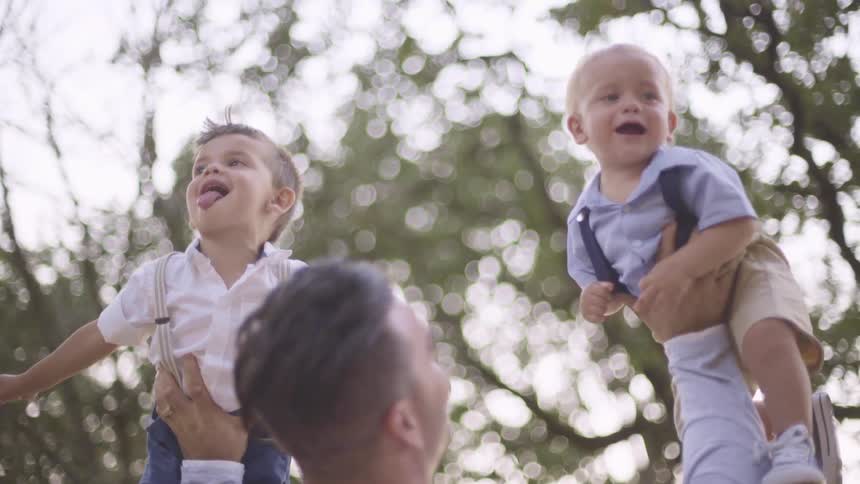 A man holds two small children up in the air while playing with them outdoors