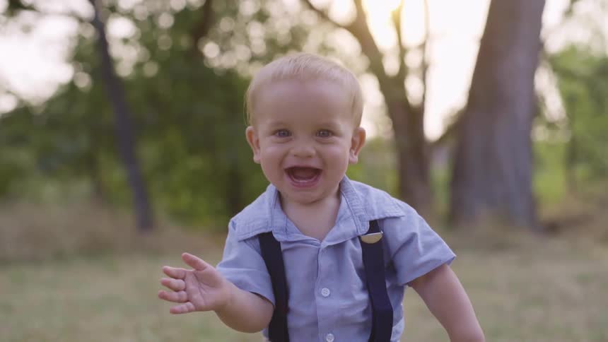 A toddler smiles and laughs happily while he stands outside on a grassy field