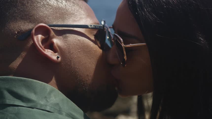 A black couple wearing sunglasses kiss standing in front of a body of water