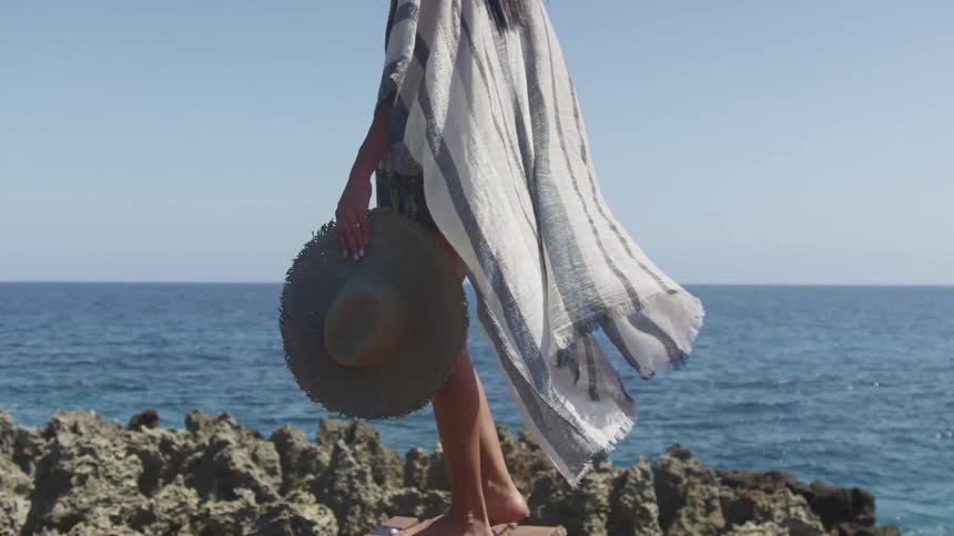 A woman stands outdoors on a sunny day while looking out upon the ocean